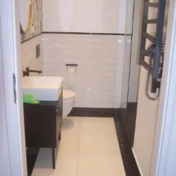 Wykończona łazienka z białymi płytkami ułożonymi w cegiełkę, czarnym pasem płytek na górze i dole ściany, umywalką na ciemnej szafce, okrągłym lustrem w złotej ramie i grzejnikiem drabinkowym...