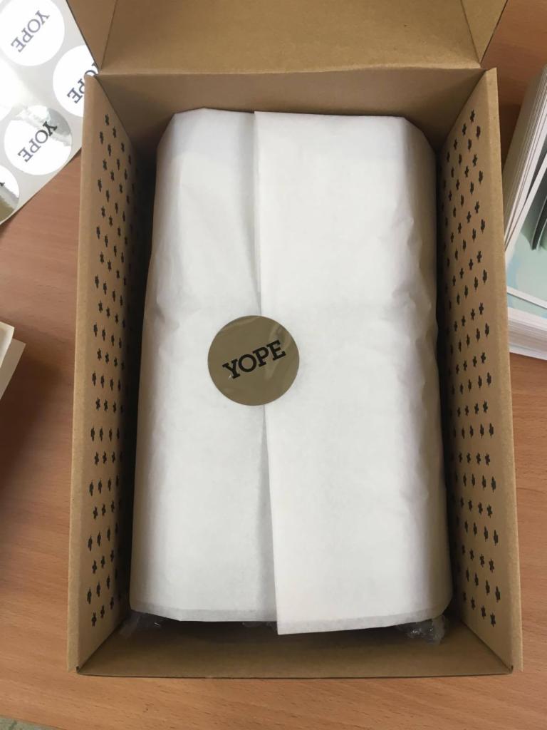 Kartonowe pudełko z zapakowanym produktem w białym papierze, zabezpieczonym okrągłą naklejką z logo firmy YOPE.