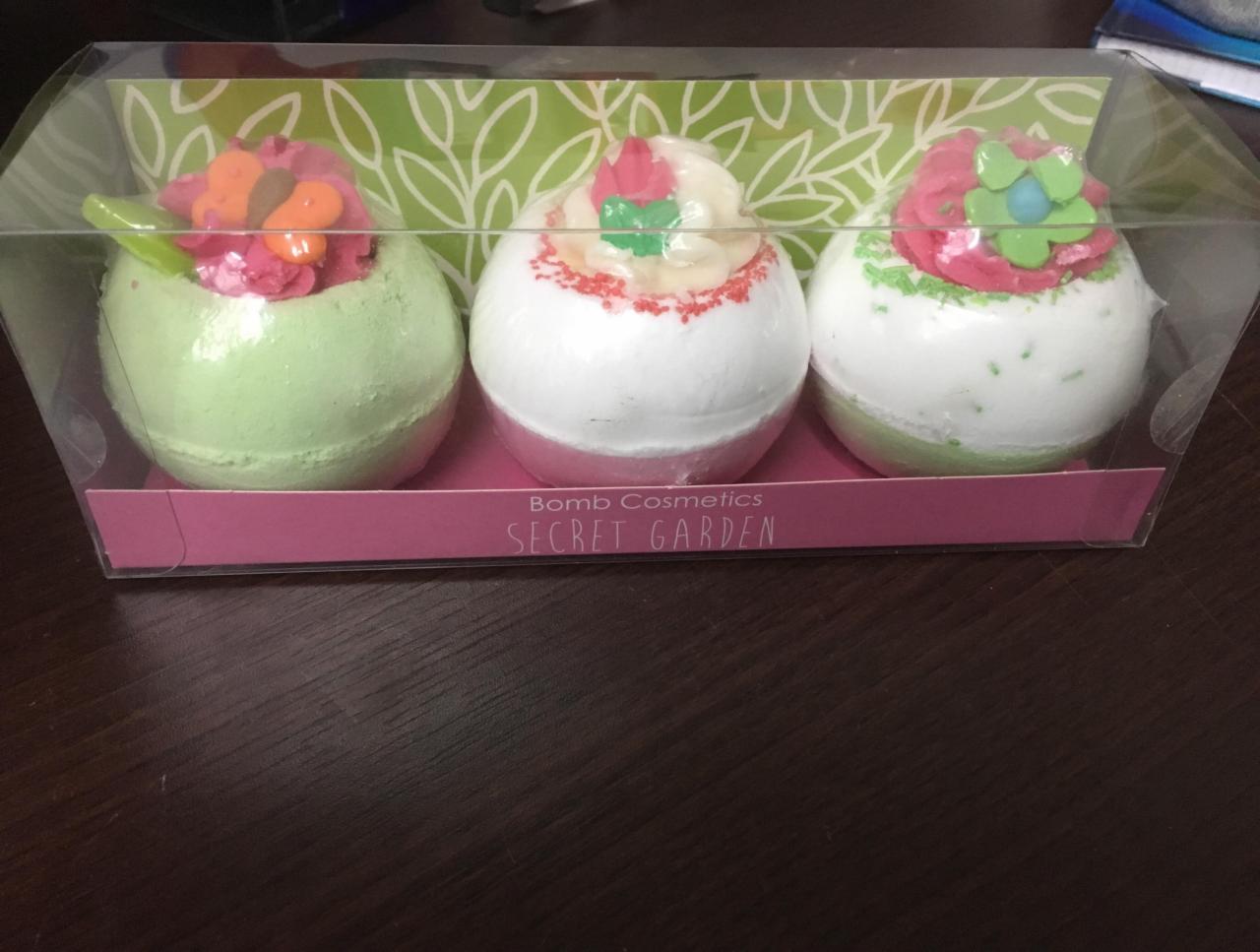 Trzy kolorowe kule do kąpieli w przezroczystym pudełku z motywem liści na tle, każda ozdobiona kremem i dekoracjami przypominającymi kwiaty.