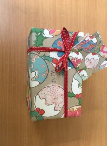 Prezent zapakowany w ozdobny papier w motywy świąteczne, przewiązany czerwoną wstążką z zawieszką, leżący na drewnianej powierzchni.