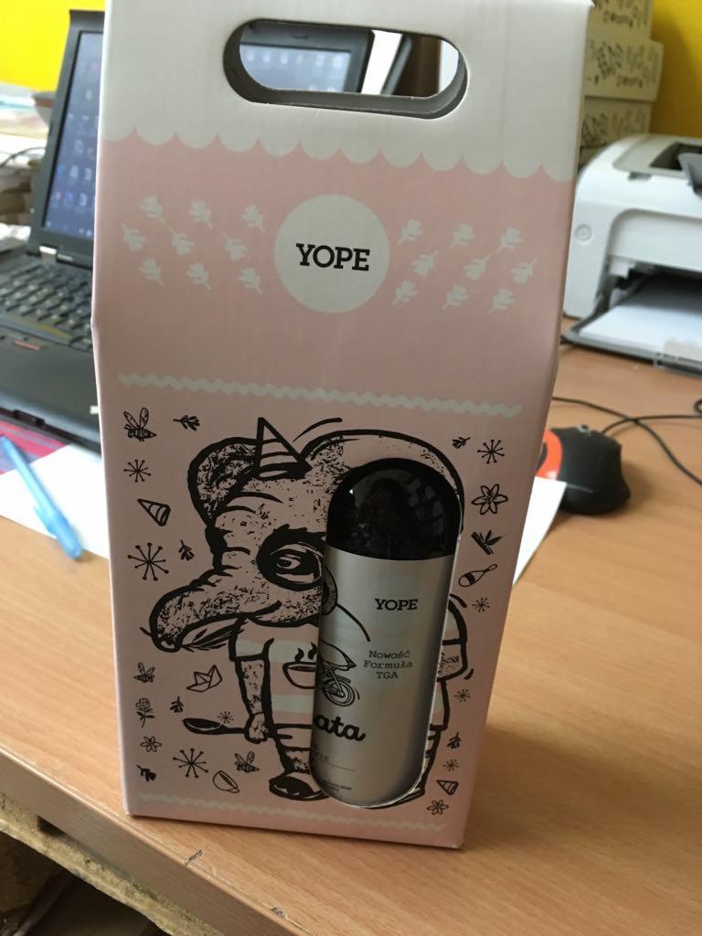 Różowy kartonowy worek z ilustracją słonia w koszulce i czapce urodzinowej, trzymającego butelkę z rozpylaczem marki YOPE, stojący na biurku z laptopem i drukarką w tle.