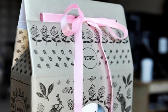 Ozdobne kartonowe pudełko z nadrukiem roślinnym i logotypem Yope, przewiązane różową satynową wstążką.