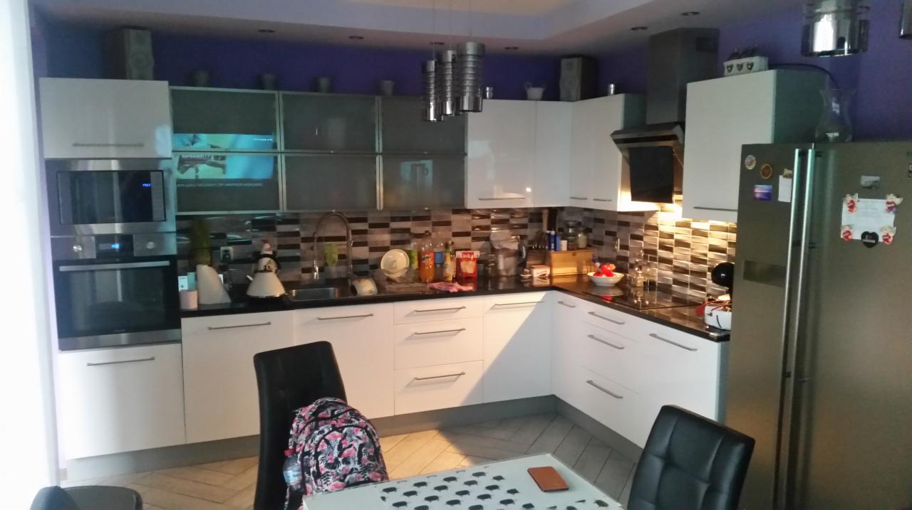 Nowoczesna, biała kuchnia z czarnym blatem i mozaikową ścianą nad blatem, widoczne sprzęty AGD i akcesoria kuchenne, fioletowa ściana w tle.