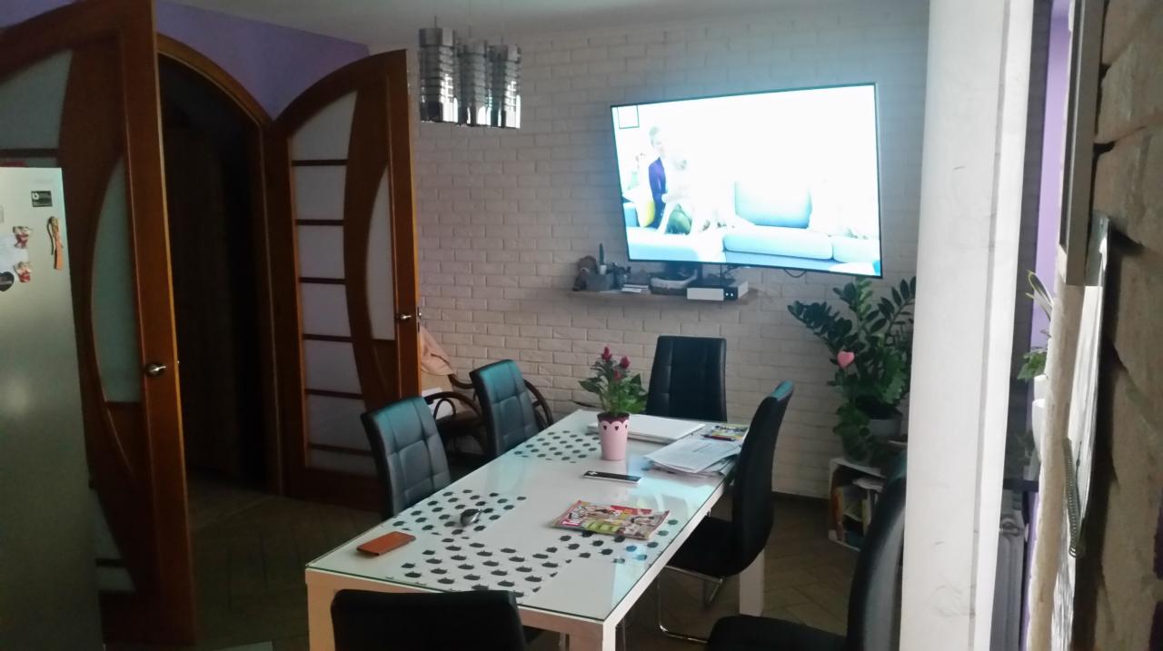 Jasna jadalnia z białym stołem, czarnymi krzesłami i telewizorem na ścianie z cegły. Widoczne drewniane drzwi z przeszkleniami i lampa wisząca.