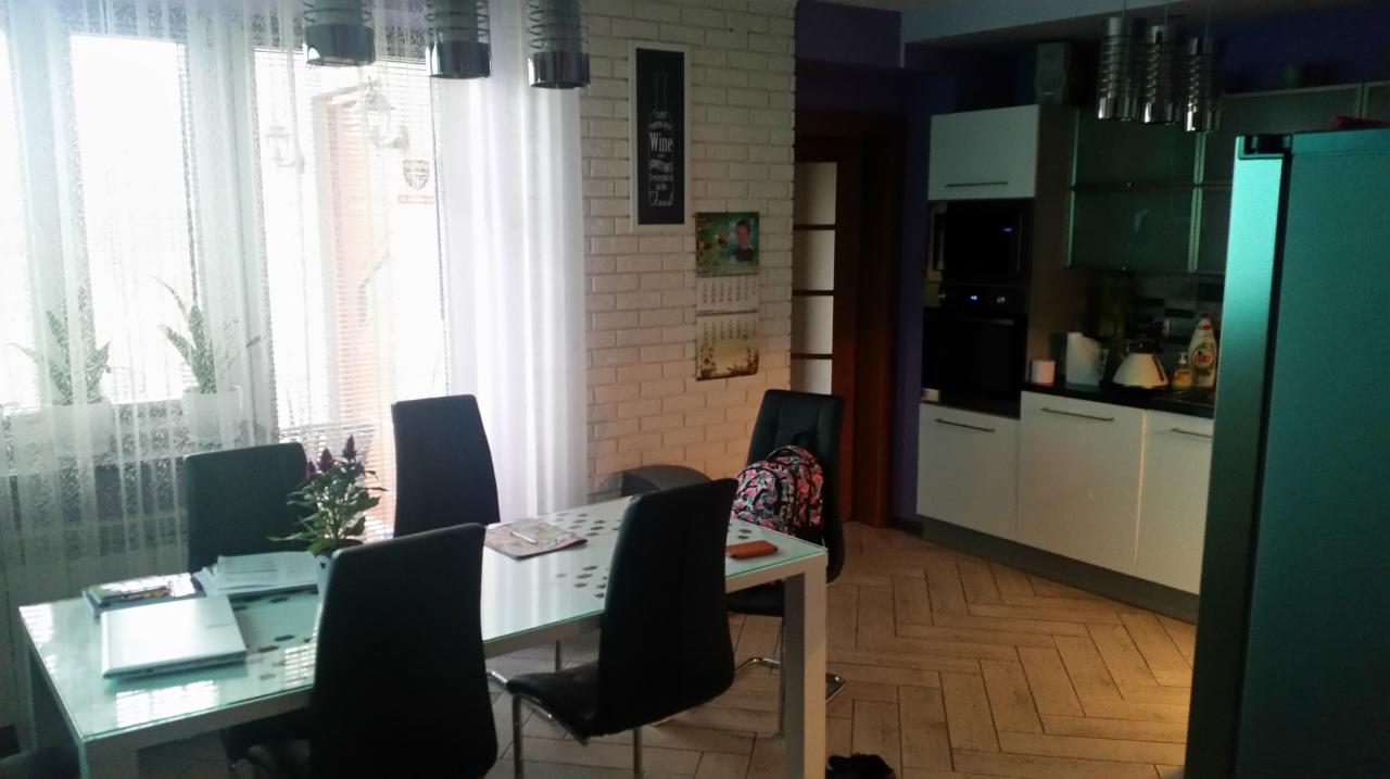 Jasna kuchnia z białymi meblami, ceglaną ścianą i szklanym stołem otoczonym czarnymi krzesłami. Na stole leży laptop i dokumenty, a przy oknie widać rośliny doniczkowe.