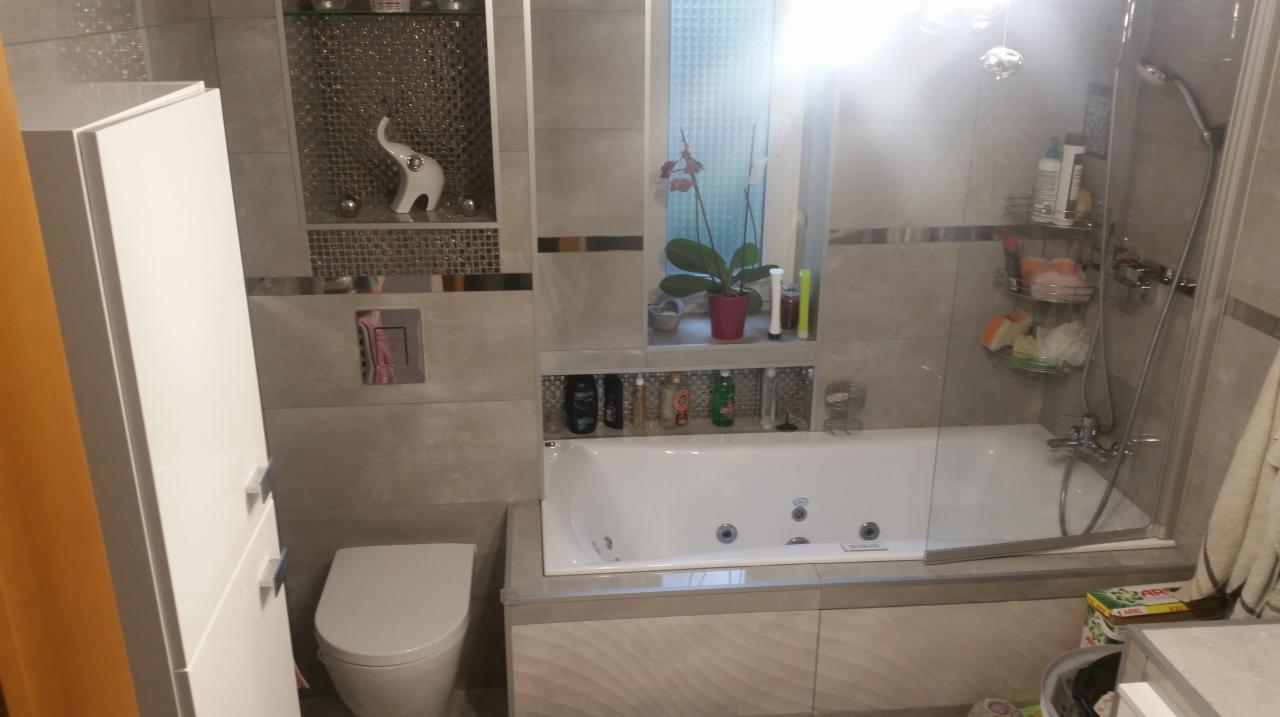 Nowoczesna łazienka z wanną z hydromasażem, szarymi płytkami, dekoracyjną mozaiką, białymi meblami i roślinami w oknie.