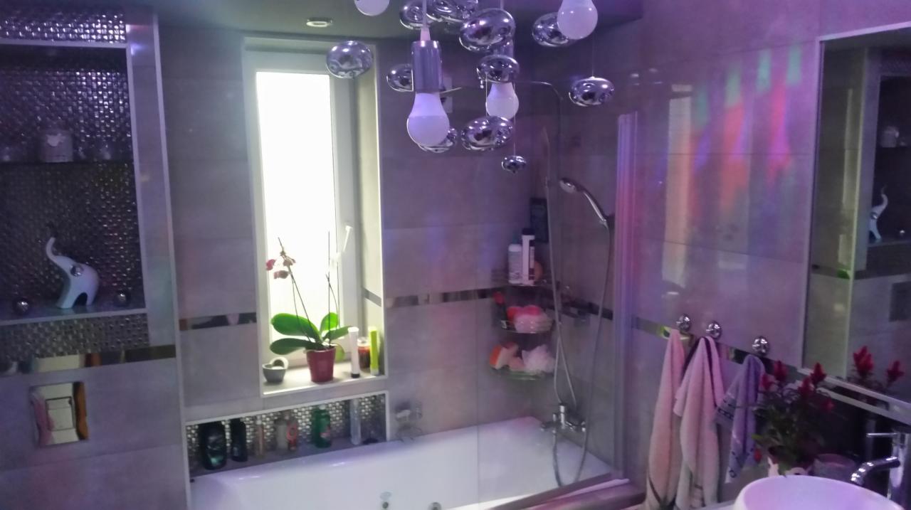 Nowoczesna łazienka z wanną, szklaną kabiną prysznicową, lustrem, dekoracyjną armaturą i oświetleniem, akcentem roślinnym w oknie oraz figurką słonia na półce.