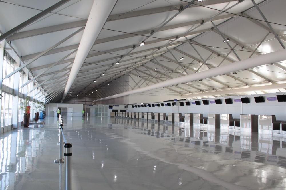 Wnętrze hali lotniskowej z widoczną stalową konstrukcją dachu, szklanymi ścianami i stanowiskami odpraw, odbijającymi się w polerowanej, szarej posadzce.
