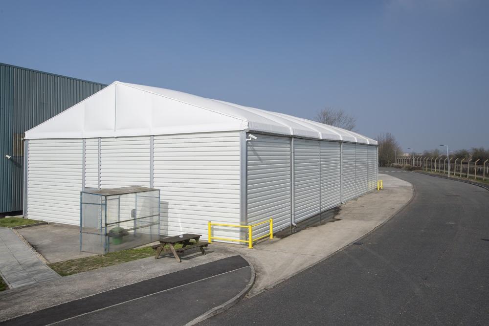 Biała hala namiotowa z panelami rolowanymi, widok z zewnątrz, obok szara hala przemysłowa, na pierwszym planie stolik piknikowy i mała szklarnia.