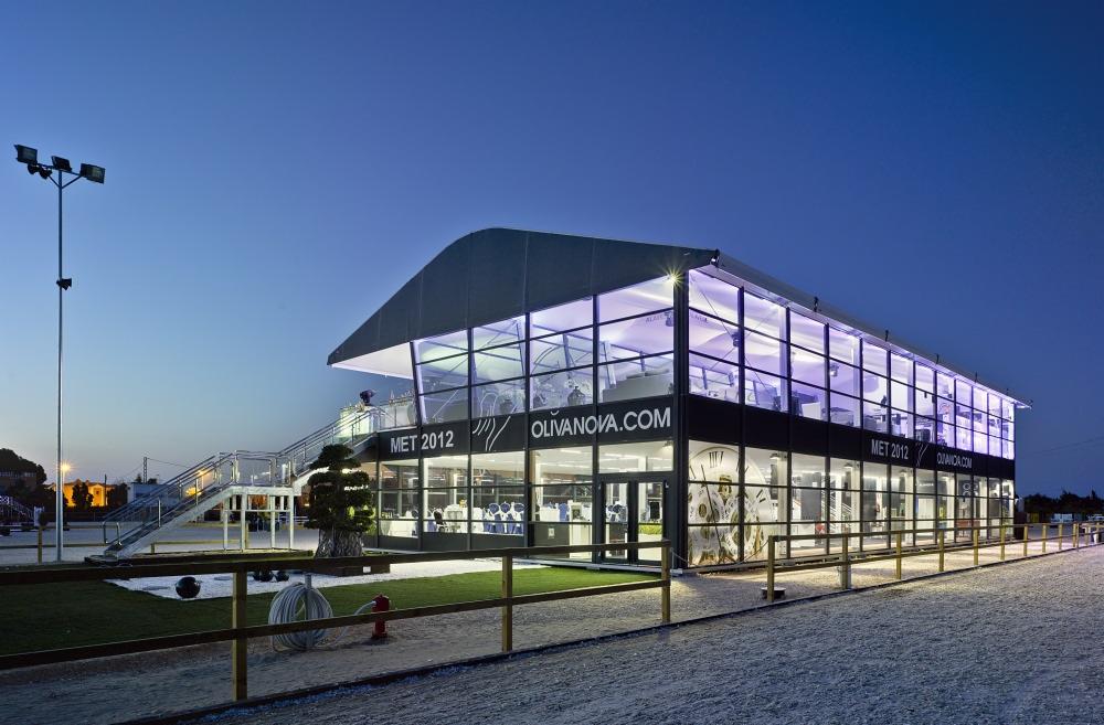 Dwukondygnacyjna hala stalowa z dużymi przeszkleniami, oświetlona od wewnątrz fioletowym światłem, z napisem OLIVANOVA.COM, widoczna z zewnątrz wczesnym wieczorem.