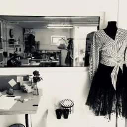 Czarno-białe zdjęcie szwalni: manekin z bluzką w paski i tiulową spódnicą, w tle pracownia z widoczną przez okno szwaczką przy maszynie, na pierwszym planie biurko z notatkami i wazonem z różą.