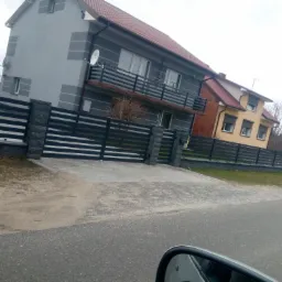 Szary dom z nową elewacją i balkonem, widoczna brama wjazdowa z szarej kostki.