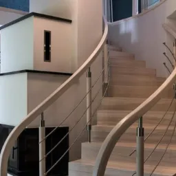 Spiralne schody z jasnego drewna z metalowymi barierkami, oświetlone przez wiszące lampy, z widocznym fragmentem zabudowy kominkowej.