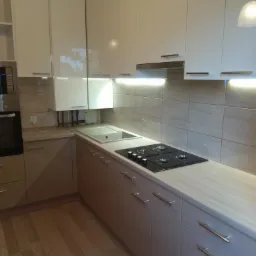 Nowoczesna, narożna zabudowa kuchenna w odcieniach beżu i bieli, z drewnianą podłogą i płytkami imitującymi kamień na ścianie, oświetlona listwami LED pod szafkami wiszącymi.