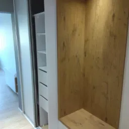 Biała szafa przesuwna z lustrem i drewnianą wstawką, widoczne półki i szuflady, na podłodze panele w jasnym odcieniu drewna.