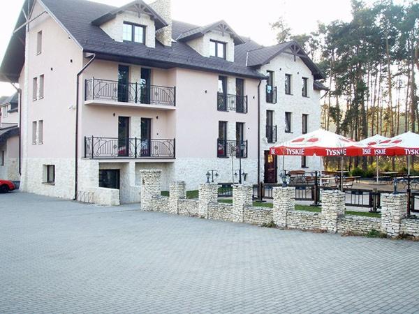 Dwupiętrowy budynek z balkonami i zewnętrznym miejscem do siedzenia pod czerwonymi parasolami Tyskie, otoczony lasem.