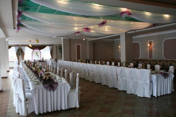 Uroczysta sala weselna z długim stołem udekorowanym białymi obrusami, fioletowymi akcentami i kwiatami, sufit ozdobiony białymi draperiami z fioletowymi wstążkami, przygotowana na przyjęcie.