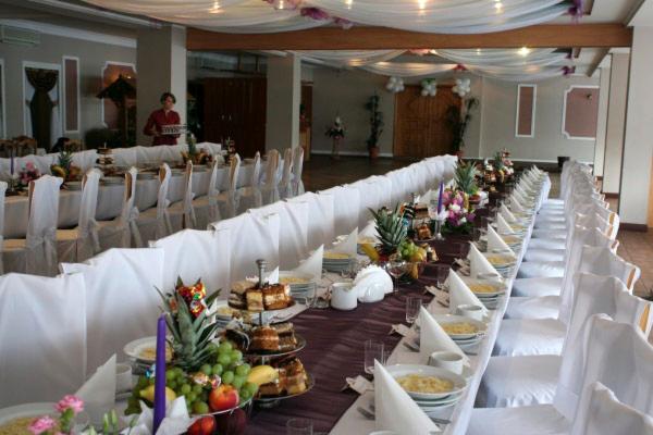 Długi stół bankietowy nakryty białym obrusem i fioletowym bieżnikiem, z dekoracjami z owoców, ciast i kwiatów, białe krzesła z pokrowcami, sala z dekoracjami sufitowymi i osobą z tacą w tle.