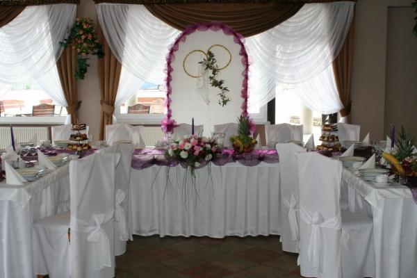 Elegancko udekorowany stół weselny z białymi pokrowcami na krzesłach, fioletowymi akcentami, kwiatami i owocami, na tle białych zasłon i dekoracji z motywem obrączek.