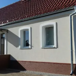 Odnowiona elewacja domu jednorodzinnego z brązową cegłą klinkierową u podstawy, kremową elewacją i nową, czerwoną dachówką ceramiczną.