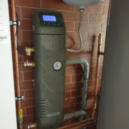 Szara jednostka centralna Hewalex z wyświetlaczem i manometrem, połączona miedzianymi rurami z instalacją w pomieszczeniu z ceglanymi ścianami.