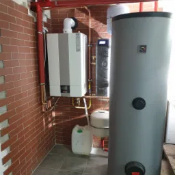 Zestaw urządzeń grzewczych w kotłowni: wiszący kocioł gazowy z szarą obudową, obok zasobnik ciepłej wody i naczynie przeponowe, całość połączona miedzianymi rurami. Na podłodze szare płytki, ściany...