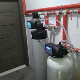 Dwa zbiorniki z elektronicznymi sterownikami połączone rurami z zaworami, instalacja w pomieszczeniu z kafelkami i drewnianymi drzwiami.