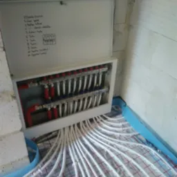 Rozdzielacz ogrzewania podłogowego w skrzynce, z widocznymi rurkami rozprowadzonymi na powierzchni z siatką, notatka z listą zadań na wewnętrznej stronie pokrywy.