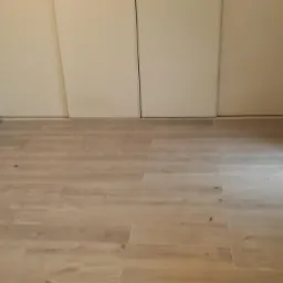 Jasne panele podłogowe ułożone w nowym mieszkaniu, widoczne białe ściany, zabudowa szafy wnękowej i okno z resztkami farby na parapecie.