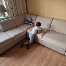 TOPMEBLE - MEBLE TAPICEROWANE ANDRZEJ ZALEWSKI - Jasny narożnik z beżową tapicerką w salonie, na którym leży pluszowy smok i bawi się dziecko z tabletem.