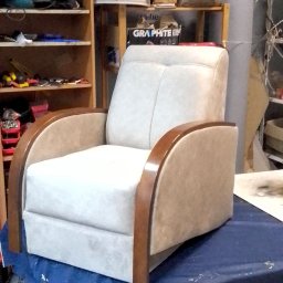 TOPMEBLE - MEBLE TAPICEROWANE ANDRZEJ ZALEWSKI - Kremowy fotel z drewnianymi podłokietnikami, stojący na stole w warsztacie tapicerskim. W tle półki z narzędziami i materiałami.