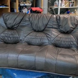 TOPMEBLE - MEBLE TAPICEROWANE ANDRZEJ ZALEWSKI - Brązowa, skórzana sofa z trzema siedziskami, z charakterystycznym pikowaniem na oparciach, stoi w warsztacie tapicerskim. Widoczne narzędzia i materiały.