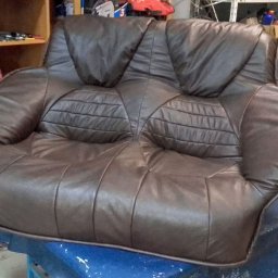 TOPMEBLE - MEBLE TAPICEROWANE ANDRZEJ ZALEWSKI - Brązowy, skórzany fotel dwuosobowy w warsztacie tapicerskim. Widoczne narzędzia i materiały, co sugeruje renowację lub produkcję. Charakterystyczne przeszycia na siedzisku.