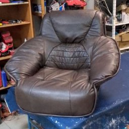 TOPMEBLE - MEBLE TAPICEROWANE ANDRZEJ ZALEWSKI - Brązowy, skórzany fotel klubowy po renowacji tapicerskiej, ustawiony na stole w warsztacie. Widoczne detale pikowania i przeszycia. W tle regały z narzędziami.