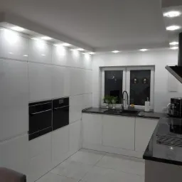 Nowoczesna kuchnia z białymi, lakierowanymi szafkami, czarnym blatem i sprzętem AGD, oświetleniem LED podszafkowym i dużym oknem.