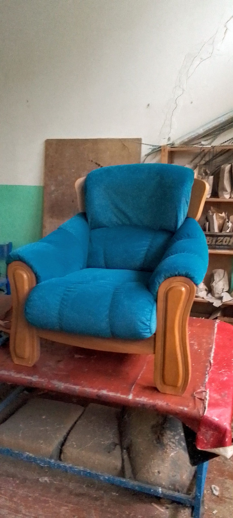 Turkusowy fotel z drewnianymi podłokietnikami na czerwonym stole w warsztacie. W tle regał z akcesoriami tapicerskimi i zielono-biała ściana.