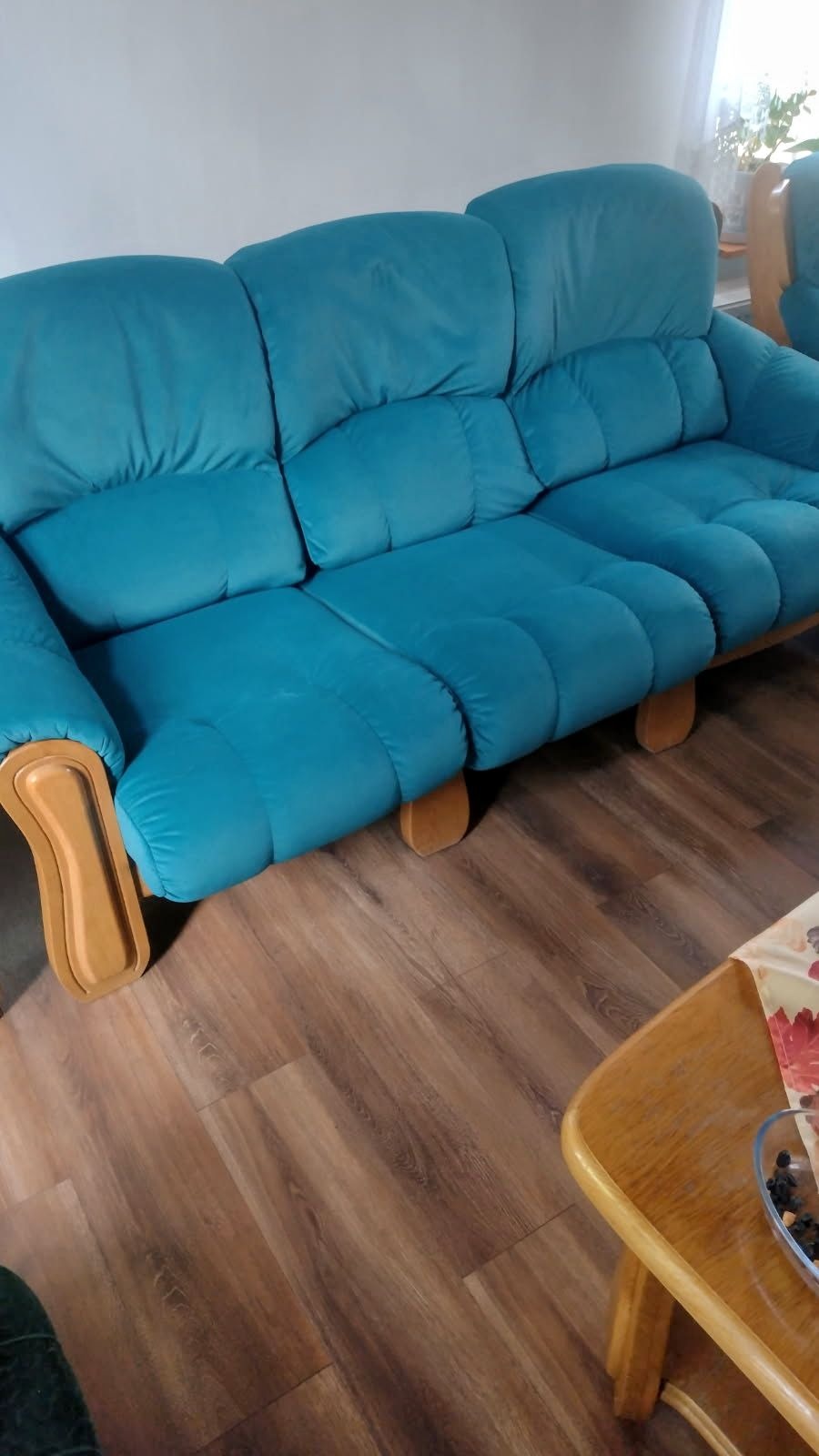 Turkusowa sofa z drewnianymi podłokietnikami na tle paneli podłogowych. Widoczny fragment stolika kawowego.