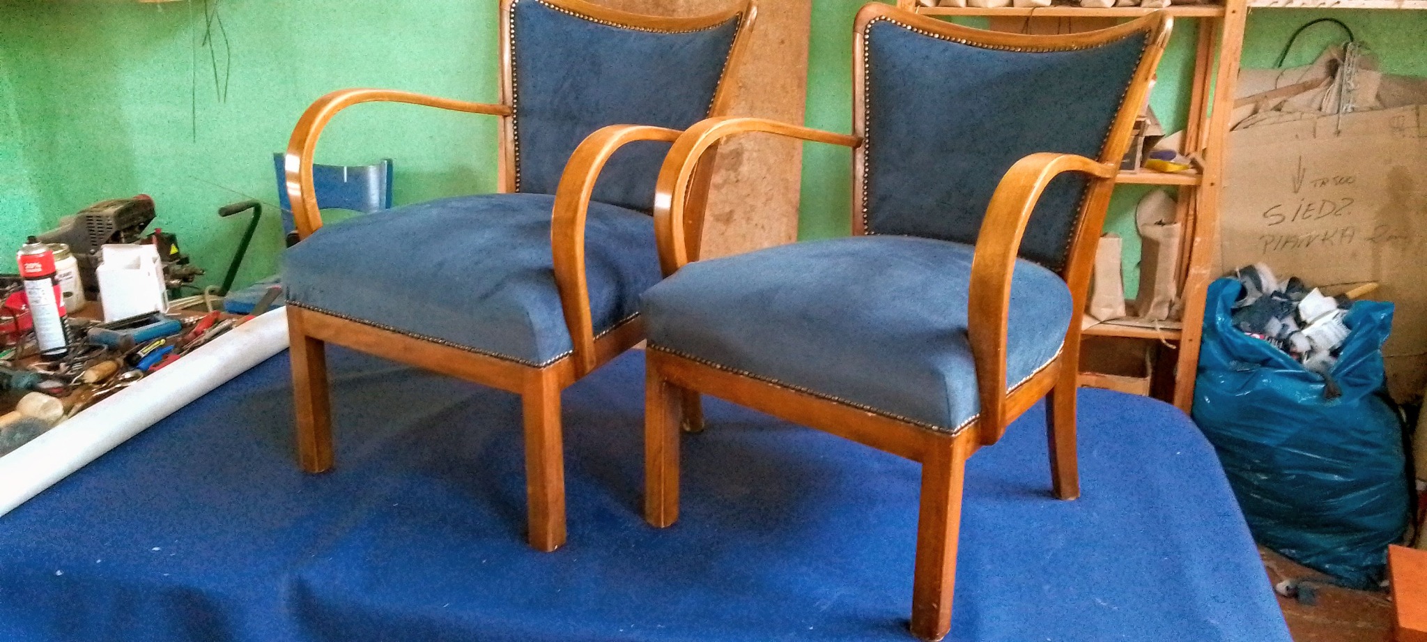 Dwa odnowione, drewniane fotele z niebieską tapicerką, ustawione w warsztacie tapicerskim. Widoczne narzędzia i materiały tapicerskie w tle.