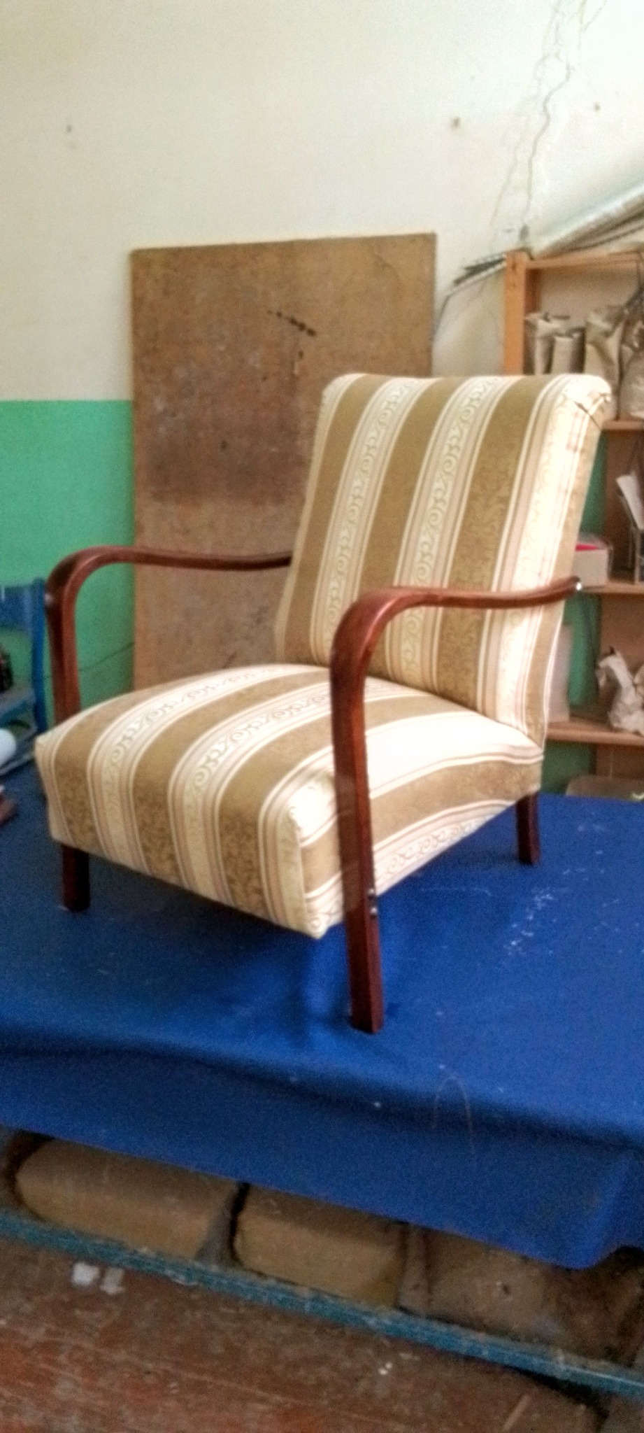 Odrestaurowany fotel z drewnianymi podłokietnikami i beżowo-białą tapicerką w paski, stojący na niebieskim materiale. Widoczne elementy warsztatu.