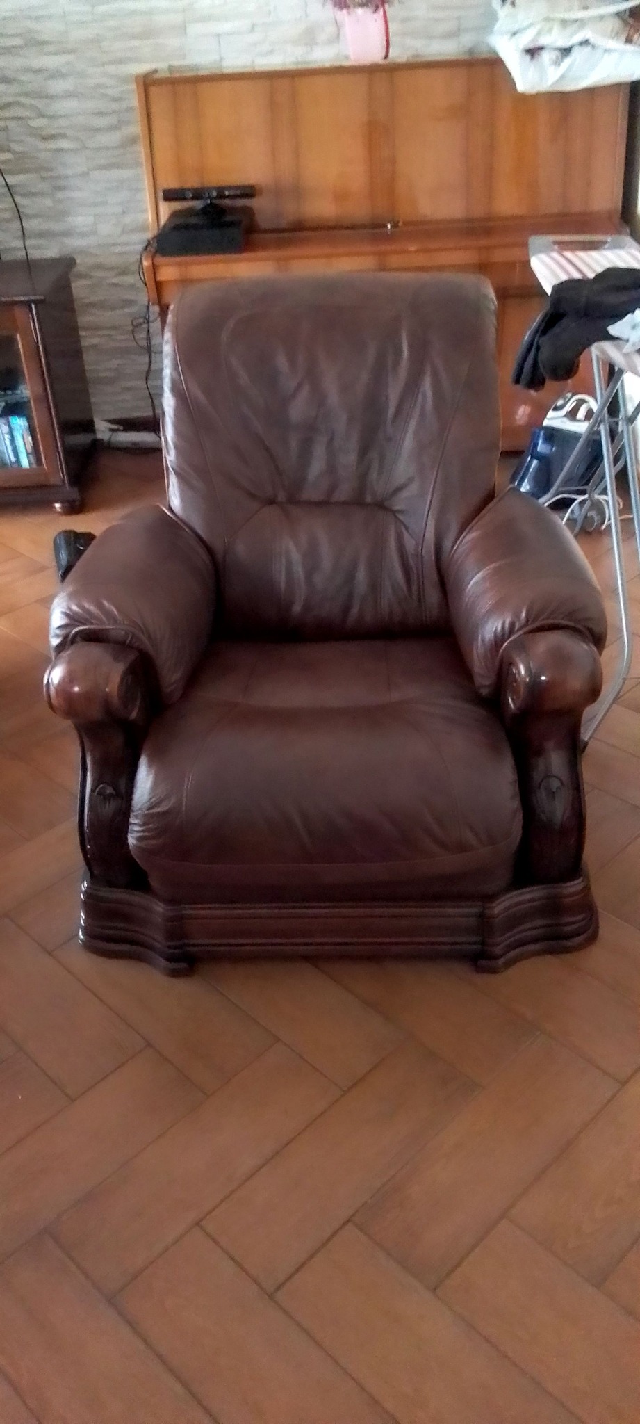 Brązowy, skórzany fotel z drewnianymi zdobieniami na podłodze z parkietu. W tle pianino i komoda. Widok z przodu, centralnie.