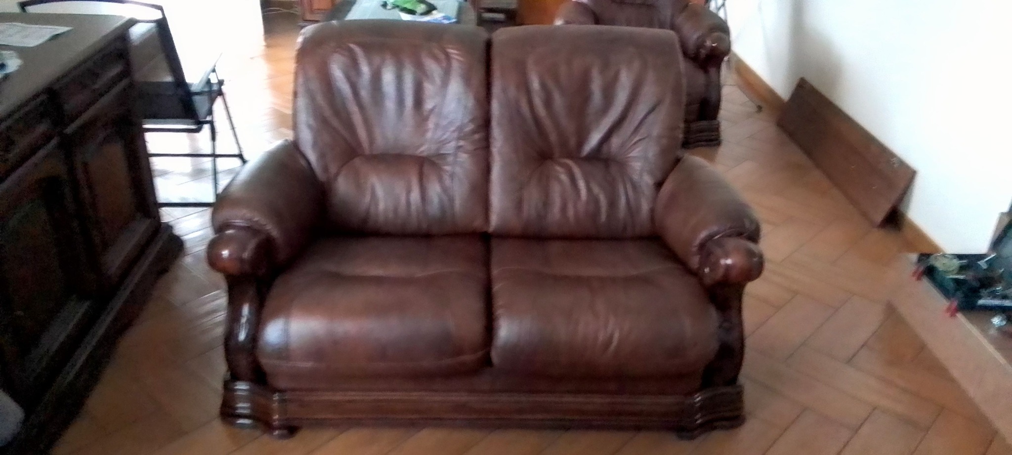 Dwuosobowa sofa ze skóry w odcieniach brązu, z widocznymi śladami użytkowania, umieszczona na parkiecie. W tle widać inne meble.