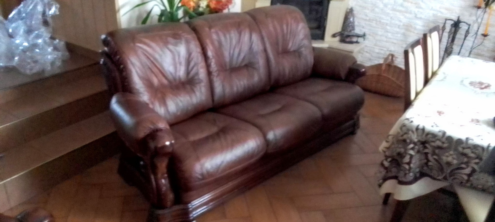 Ciemnobrązowa, skórzana sofa trzyosobowa z dekoracyjnymi drewnianymi elementami, ustawiona w jasnym salonie z parkietem i widocznym fragmentem stołu.