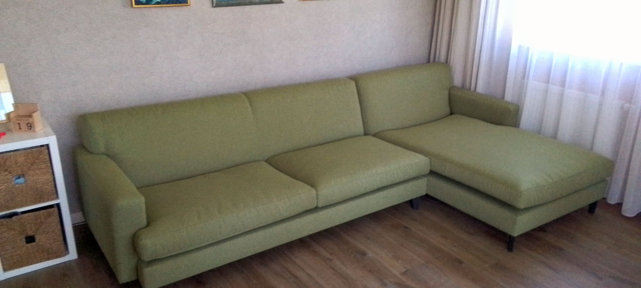 Jasnozielona, tapicerowana sofa narożna z leżanką, ustawiona na drewnianej podłodze, oświetlona naturalnym światłem z okna.