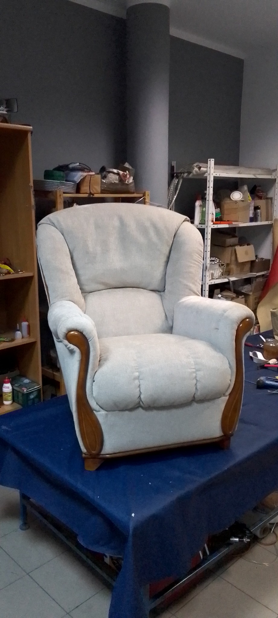 Beżowy fotel z drewnianymi podłokietnikami stoi na niebieskim materiale w warsztacie tapicerskim; w tle półki z narzędziami.