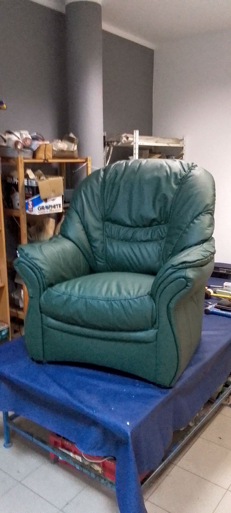 Zielony, skórzany fotel klubowy na niebieskim stole w warsztacie tapicerskim; widoczne narzędzia i półki z materiałami.