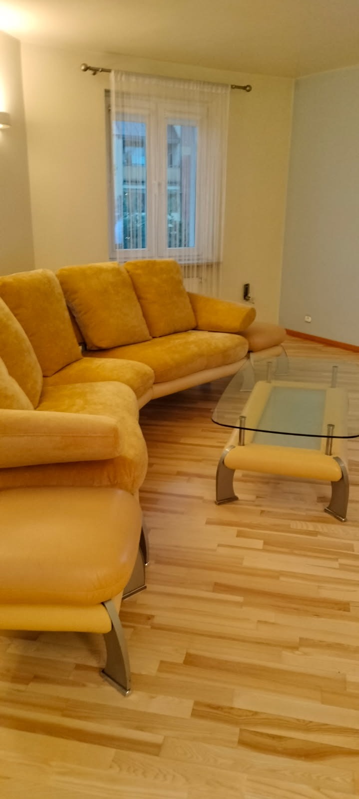 Żółta, tapicerowana sofa narożna z niskim stolikiem kawowym na drewnianej podłodze w jasnym salonie z oknem z białymi firankami. Widok z perspektywy.