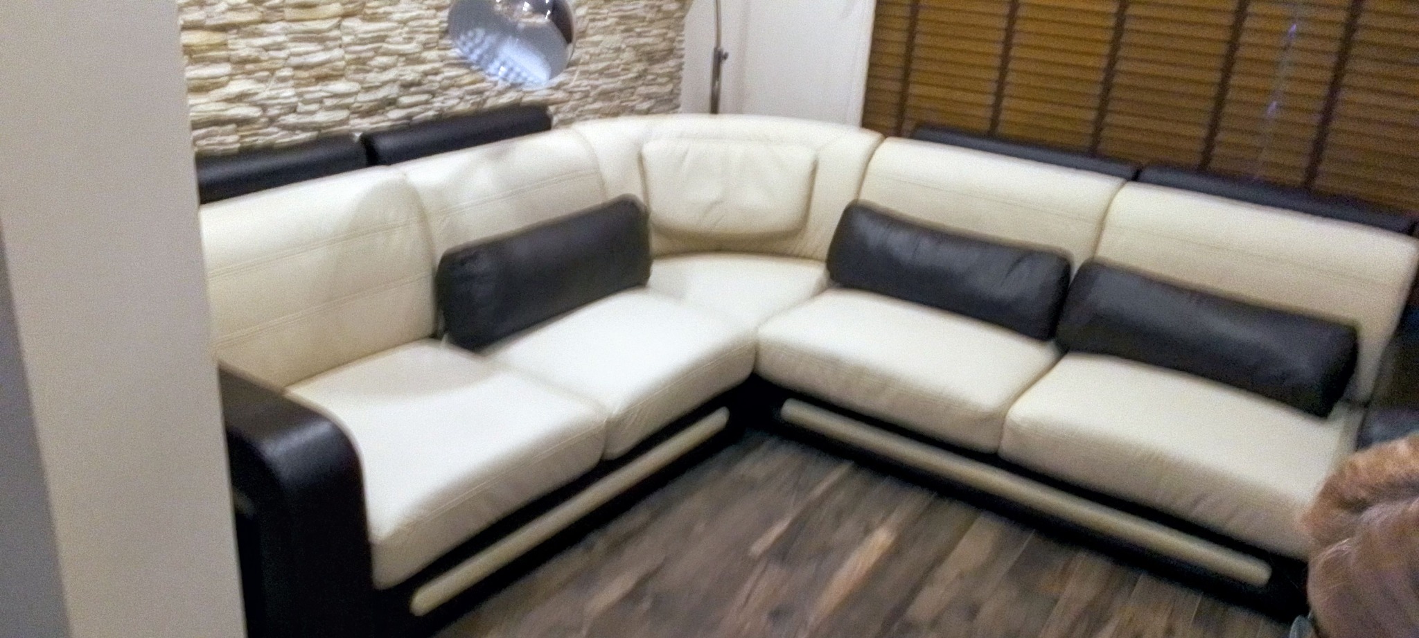 Narożnik tapicerowany w odcieniach bieli i czerni, z podłużnymi poduszkami, na tle ściany z imitacji kamienia i drewnianych żaluzji. Widok z perspektywy podłogi.