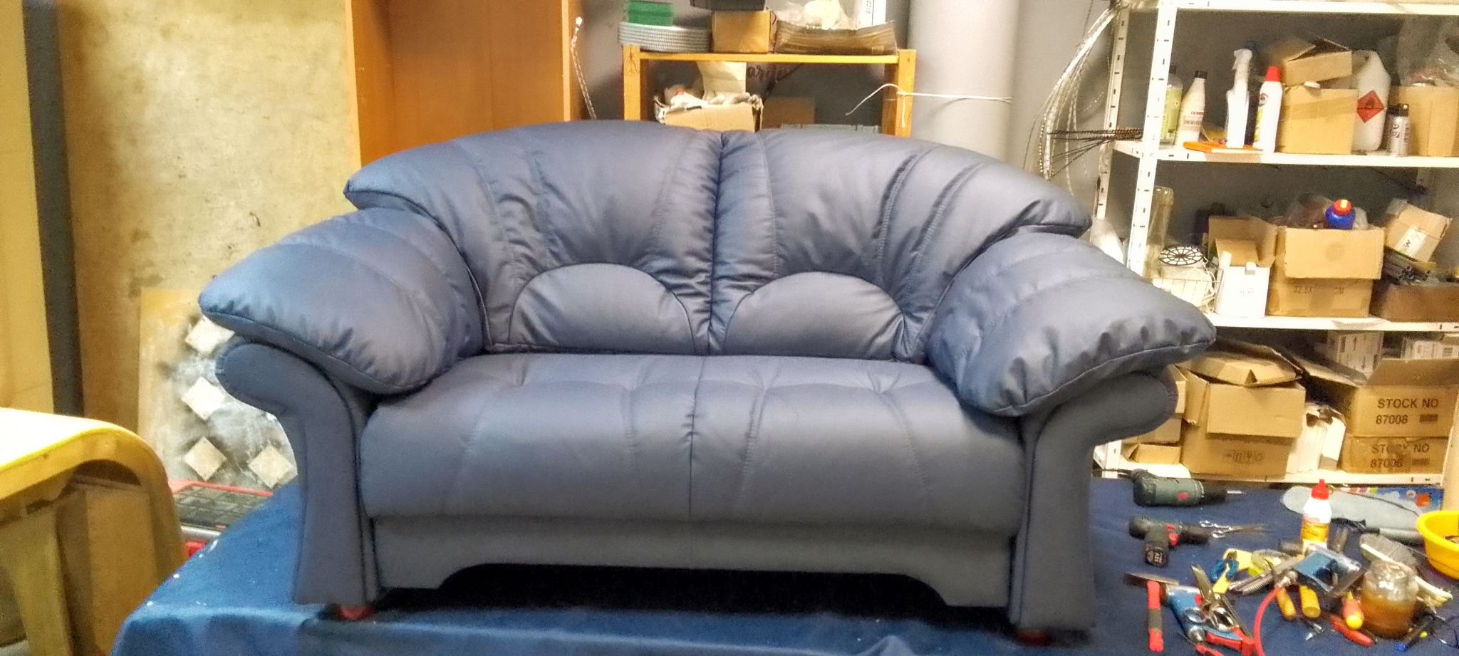 Niebieska sofa dwuosobowa z pikowanymi siedziskami i oparciem, widoczna w warsztacie tapicerskim z narzędziami na stole i regałami w tle. Charakterystyczne zaokrąglone podłokietniki.