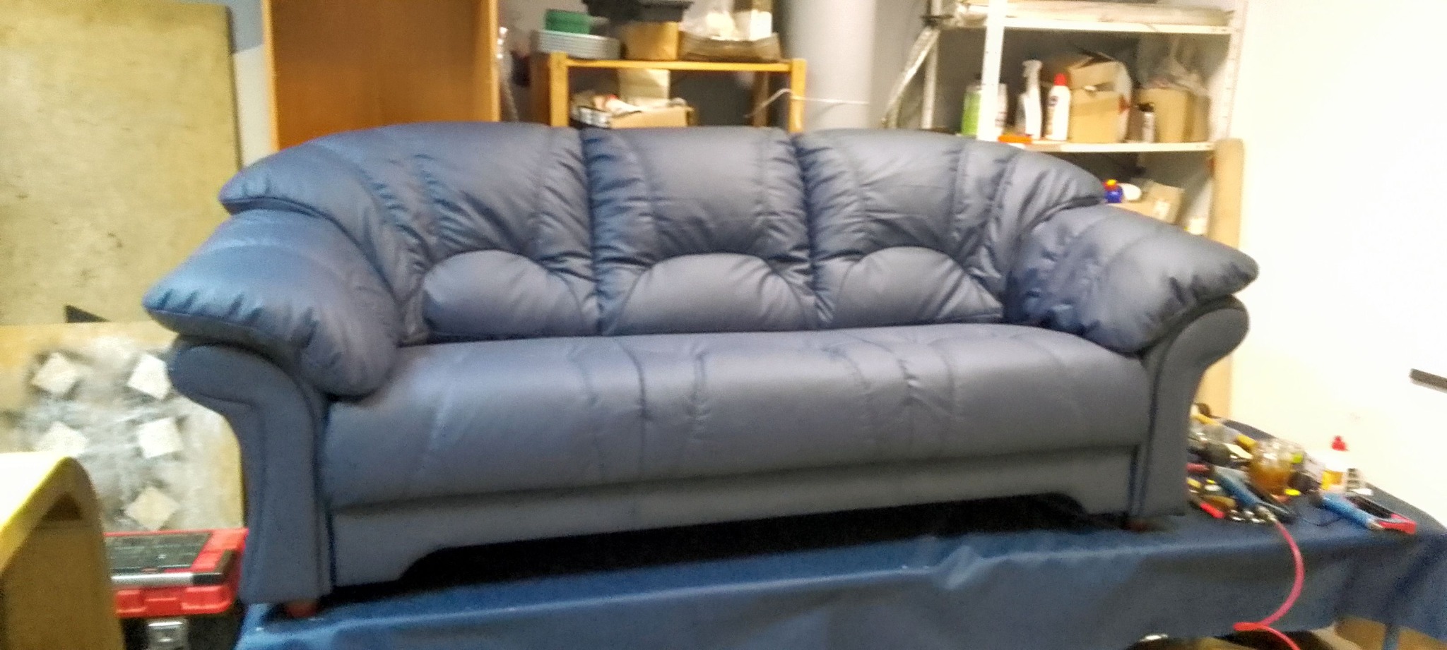 Niebieska sofa z charakterystycznym pikowaniem na siedzisku i oparciu, ustawiona w warsztacie tapicerskim z widocznymi narzędziami i materiałami w tle.