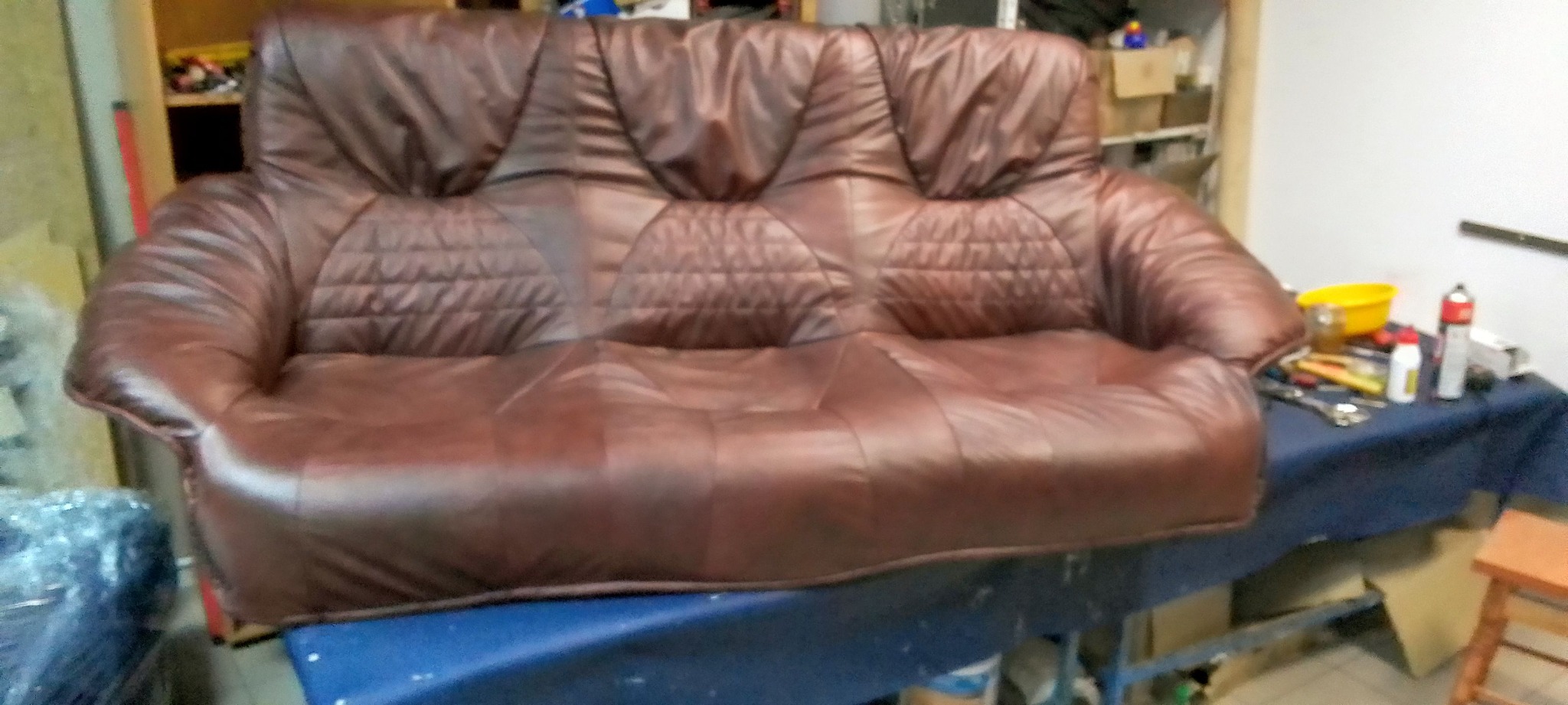 Brązowa, skórzana sofa z pikowanymi elementami, ustawiona na stole w warsztacie tapicerskim. Widoczne narzędzia i akcesoria, sugerujące proces renowacji lub tworzenia.